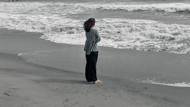 Mulher sozinha de pé na praia diante do mar agitado, sob céu nublado